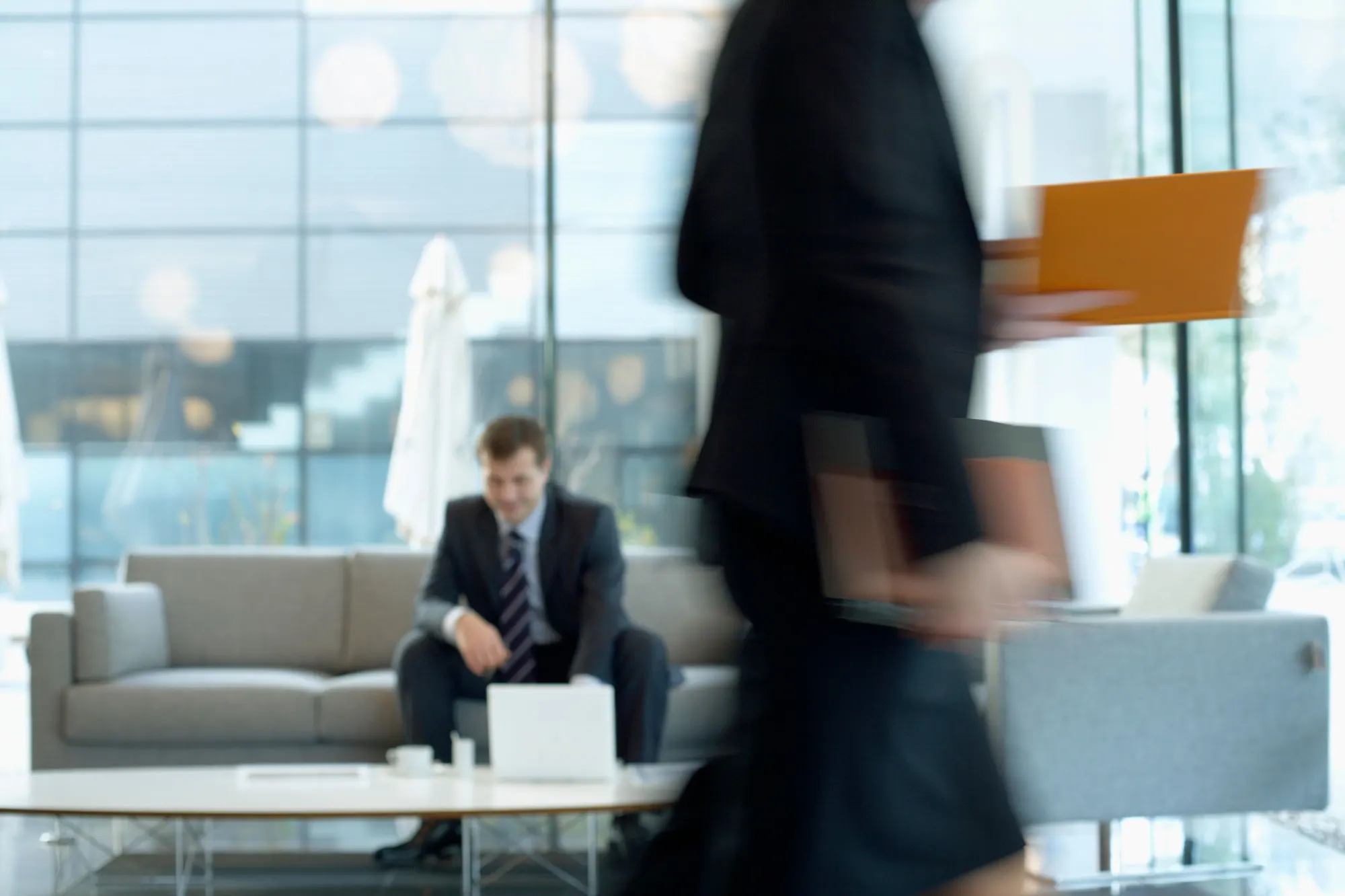 A visitor waiting in an office lobby as a colleague walks past with documents